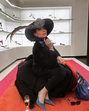 Mulher posa com elegância em loja de sapatos, usando chapéu vistoso e vestido preto