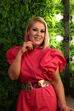 Mulher sorri com vestido rosa e cinto dourado, exibindo brincos e anel a condizer