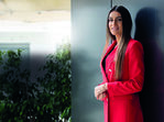 Mulher de blazer vermelho posa junto a uma parede escura e uma planta