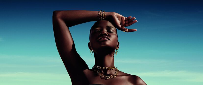Mulher com acessórios dourados e pele escura posa sob o céu azul