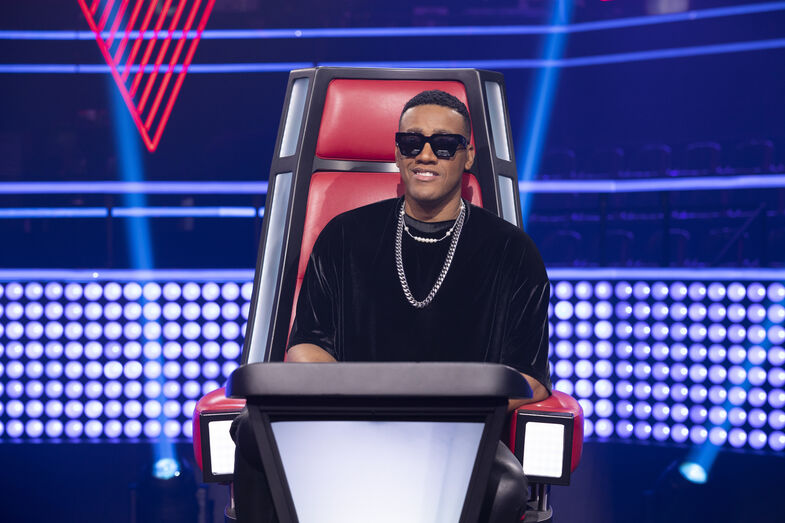 Homem sorridente, de óculos de sol, está sentado na cadeira vermelha do The Voice