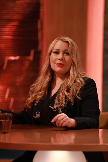 Mulher loira com batom vermelho posa para foto sentada a uma mesa