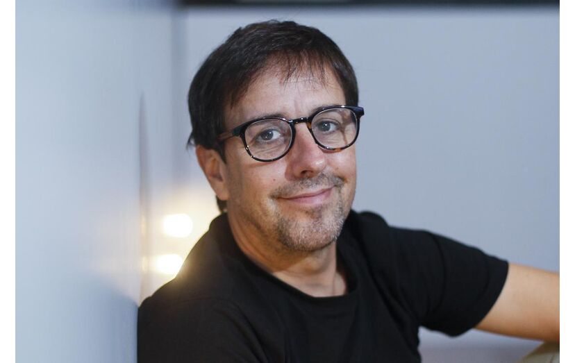 Homem sorridente, de óculos, encostado a uma parede