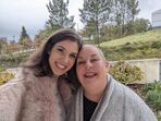 Duas mulheres sorriem juntas ao ar livre, com árvores e vegetação no fundo.
