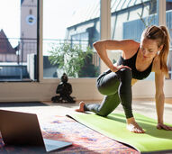 Mulher pratica exercício em casa, com tapete de ioga e computador portátil por perto