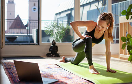 Mulher pratica exercício em casa, com tapete de ioga e computador portátil por perto