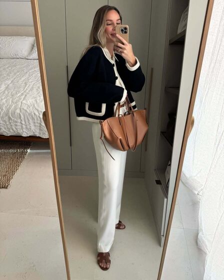 Mulher tira selfie em frente ao espelho com casaco preto, calças brancas e mala castanha