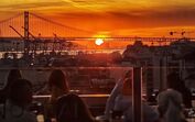 Pessoas desfrutam de um pôr do sol numa esplanada com vista para a ponte de Lisboa