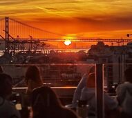 Pessoas desfrutam de um pôr do sol numa esplanada com vista para a ponte de Lisboa