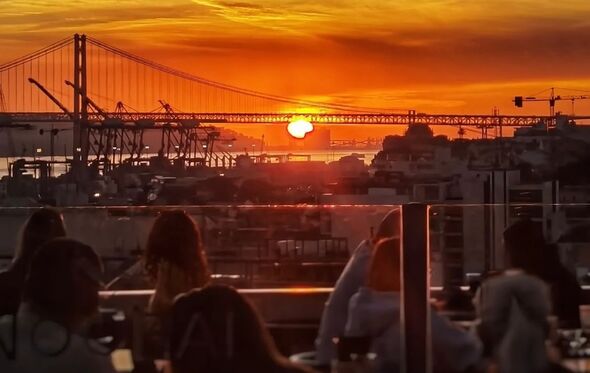 Pessoas desfrutam de um pôr do sol numa esplanada com vista para a ponte de Lisboa