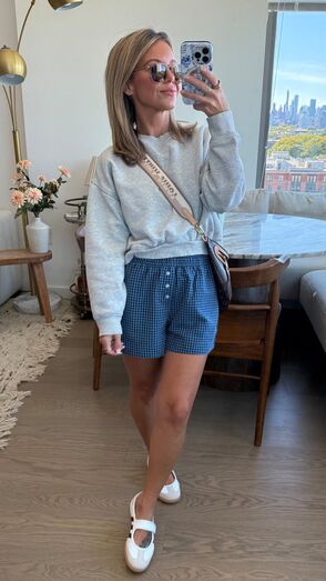 Mulher posa com calções de padrão axadrezado e camisola cinzenta num apartamento com vista para a cidade