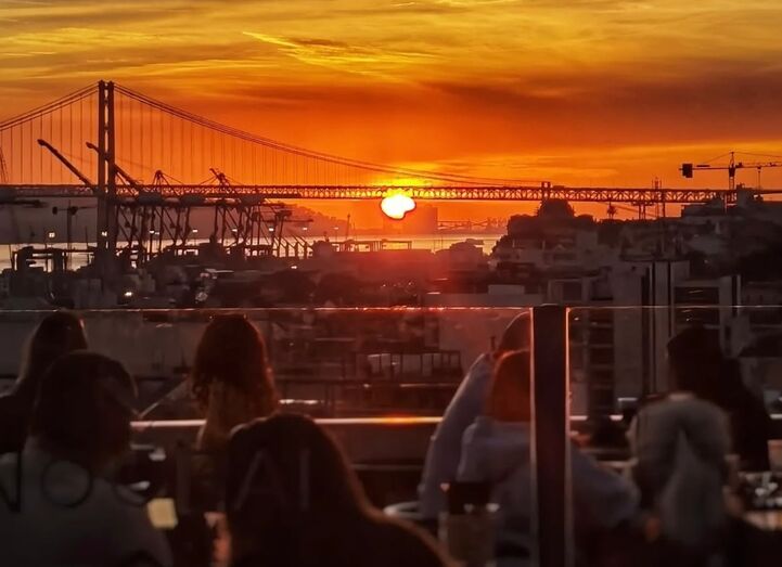 Pessoas desfrutam de um pôr do sol numa esplanada com vista para a ponte de Lisboa