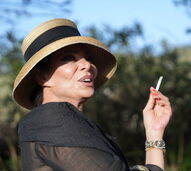 Mulher com chapéu de palha fuma cigarro com pulseiras douradas