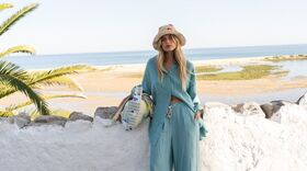 Modelo com chapéu de palha e conjunto de calças e camisa azul turquesa, junto ao mar