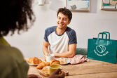 Um homem sorri com pães e café perto de um saco Too Good To Go