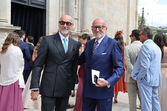 Dois homens em fatos elegantes posam sorridentes em frente a uma igreja